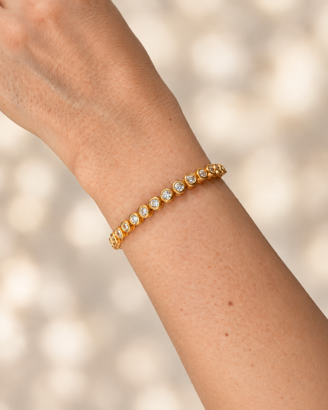 A gold tennis bracelet with small clear gemstones worn on a person's wrist, shown against a softly blurred light beige background.