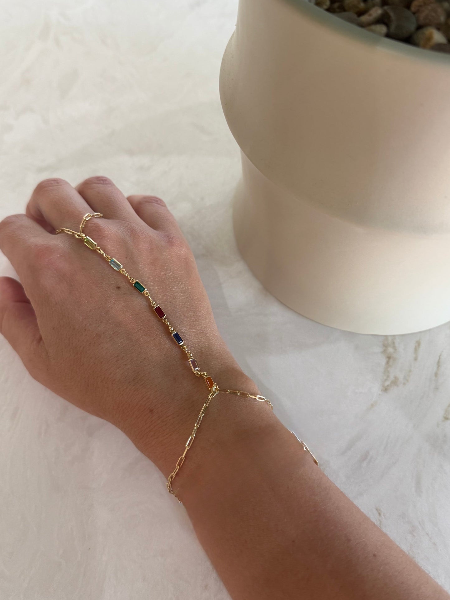 A delicate gold chain hand bracelet with small colorful gemstone accents is shown worn on a person's hand, resting on a white textured surface next to a potted plant.
