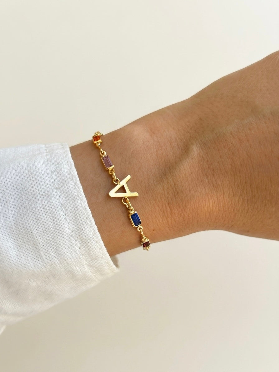 A gold initial bracelet with a letter 'A' charm, accented with small purple and blue beads, shown on a person's wrist wearing a white sleeve.
