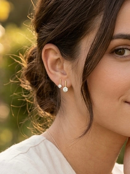 A close-up of a woman's ear wearing a pair of small gold hoop earrings with clear round gemstones, set against a softly blurred outdoor background.