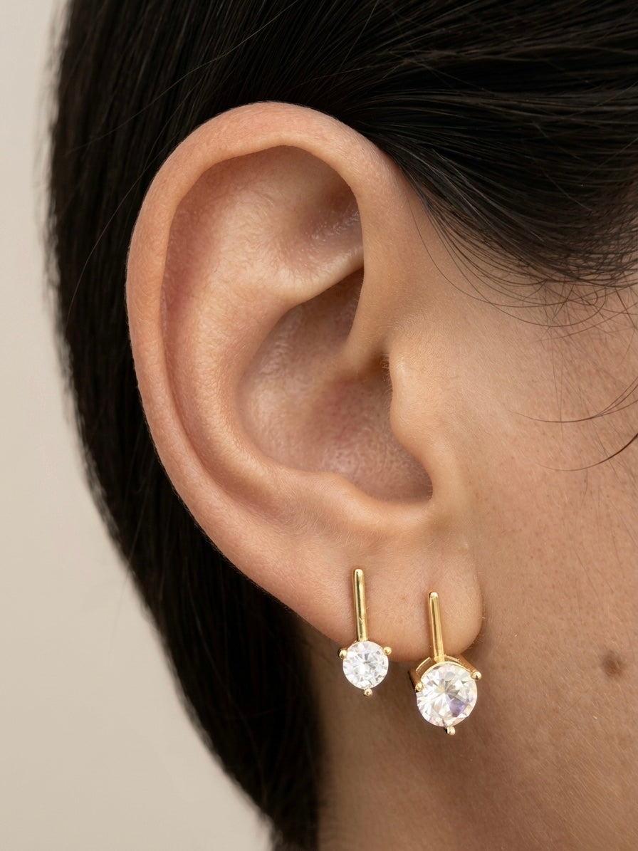 A close-up of a person's ear wearing a pair of small gold stud earrings with clear round gemstones, against a plain light beige background.