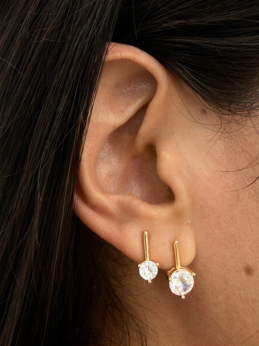 A close-up of a person's ear wearing a pair of gold stud earrings with round clear gemstones.