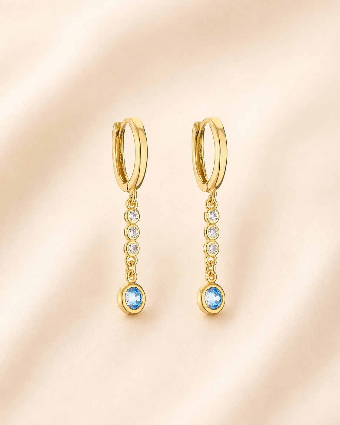 A pair of gold huggie hoop earrings with a dangling vertical bar set with clear cubic zirconia stones and a round light blue gemstone at the bottom, displayed on a soft beige fabric background.