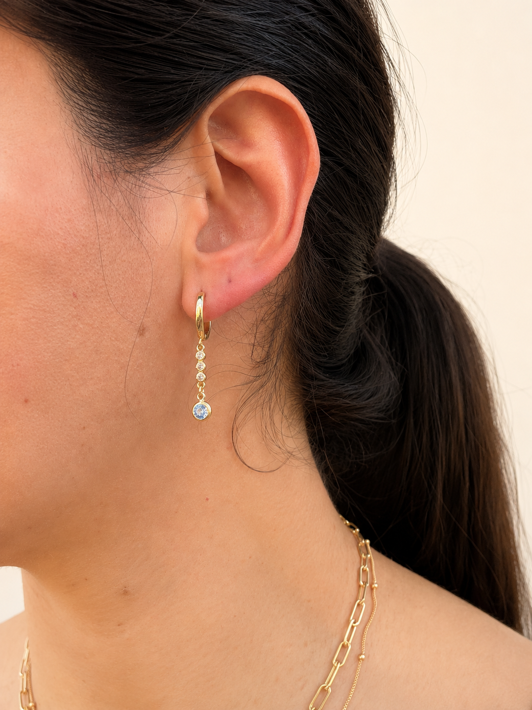A close-up side profile of a woman wearing a delicate gold huggie hoop earring with a small drop of clear stones ending in a round light blue stone, against a warm cream background.