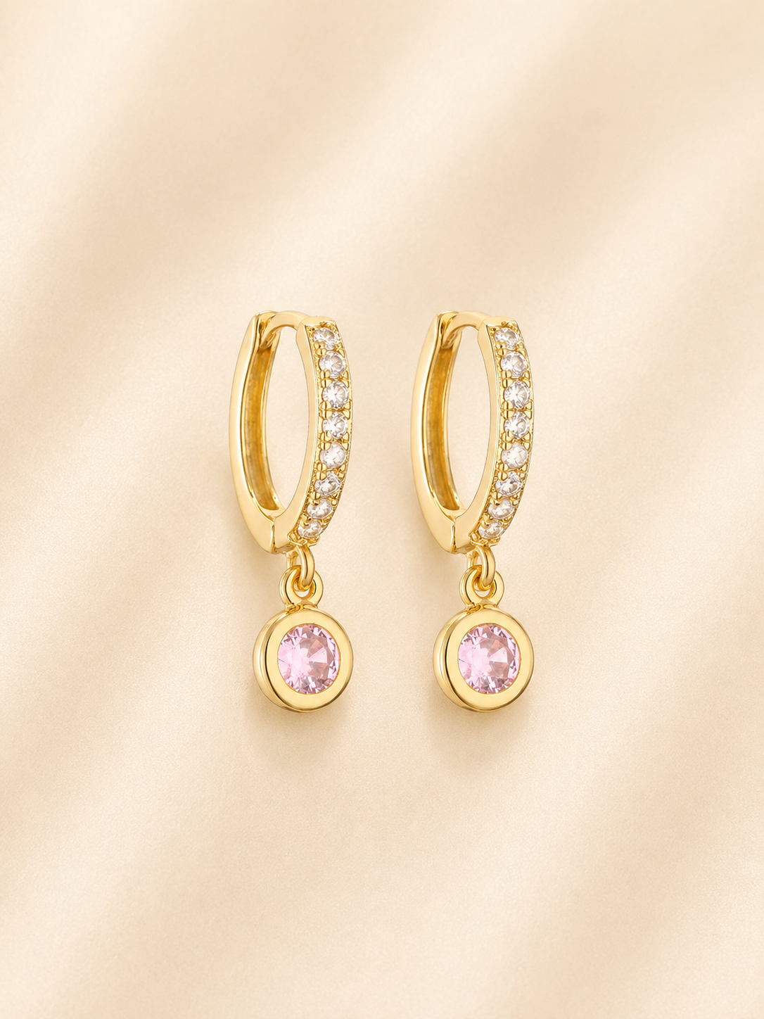 A pair of gold huggie hoop earrings with pavé-set clear stones and a dangling round pink gemstone, displayed on a soft beige fabric background.