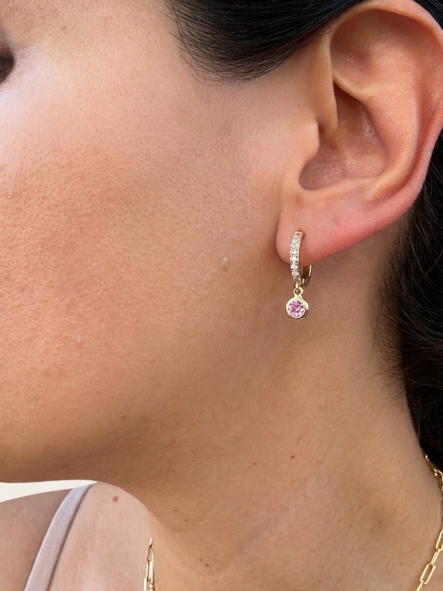 A close-up of a model's ear wearing a delicate gold huggie hoop earring adorned with small clear stones and a dangling pink gemstone charm.