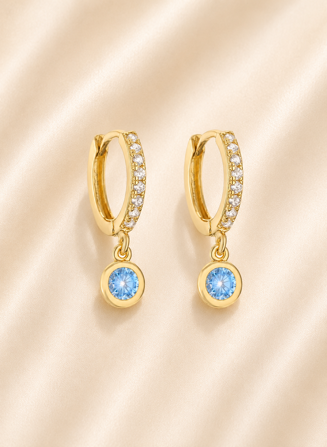 A pair of gold huggie hoop earrings with small clear stones along the top band and a dangling round light blue gemstone at the bottom, displayed on a soft beige satin fabric background.