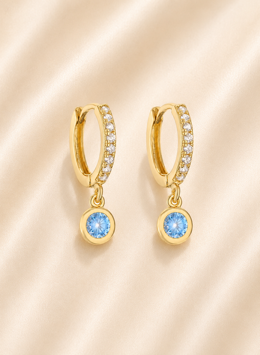 A pair of gold huggie hoop earrings with small clear stones along the top band and a dangling round light blue gemstone at the bottom, displayed on a soft beige satin fabric background.