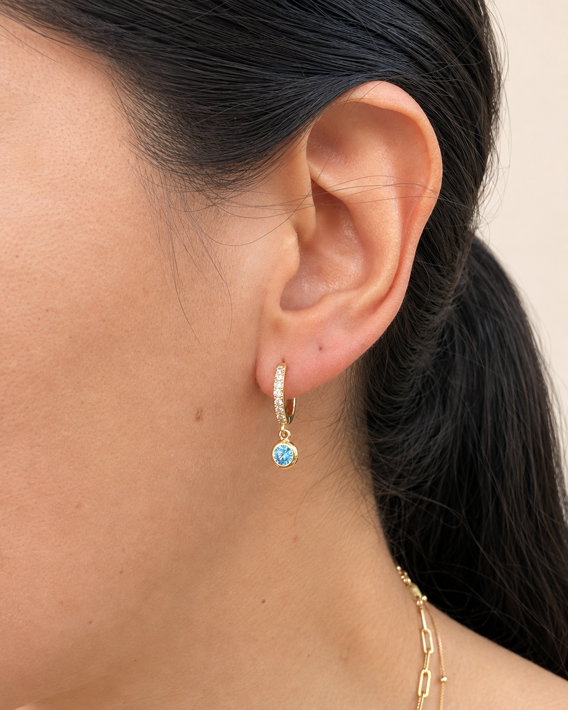 A close-up of a model's ear wearing a delicate gold huggie hoop earring with a small round light blue gemstone and tiny clear stones along the top.