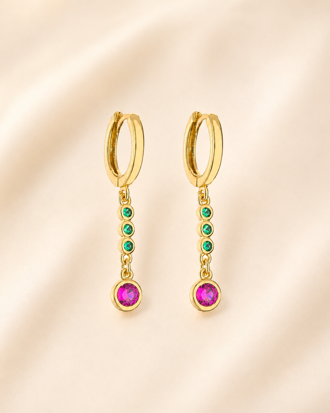 A pair of gold huggie hoop earrings with dangling chains, featuring small green gemstones and a larger dark pink gemstone at the bottom, displayed on a soft beige fabric background.