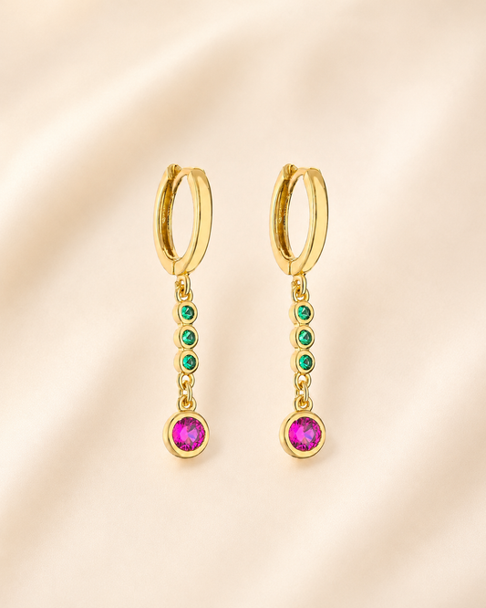 A pair of gold huggie hoop earrings with dangling chains, featuring small green gemstones and a larger dark pink gemstone at the bottom, displayed on a soft beige fabric background.
