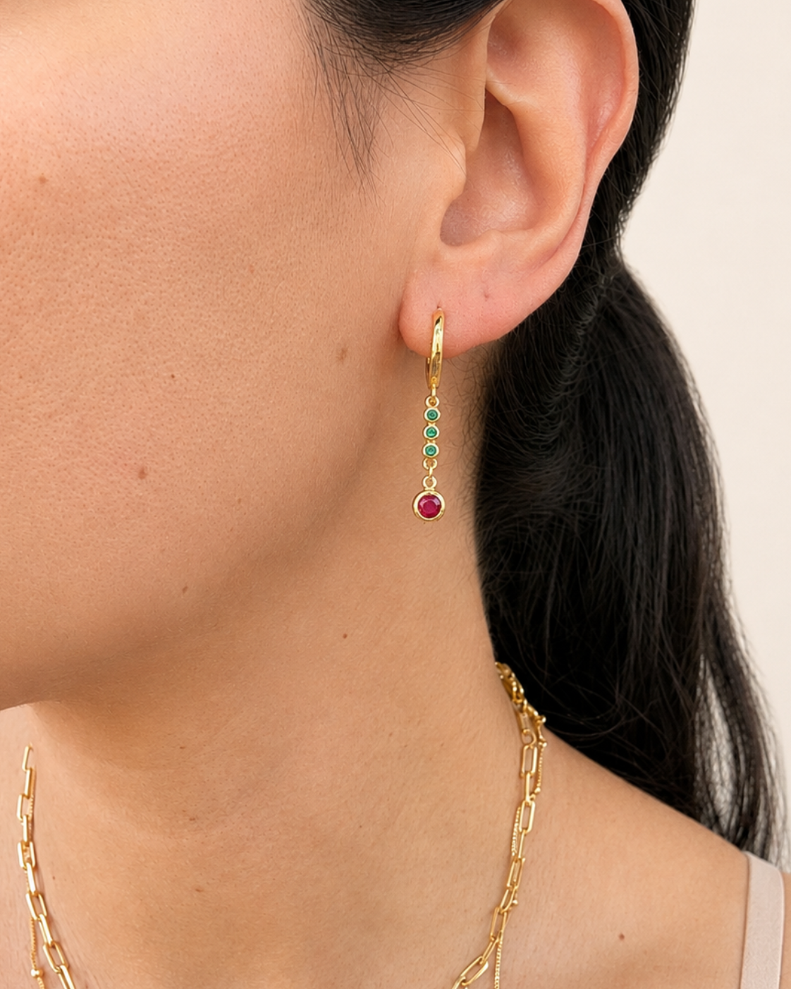 A close-up of a model's ear wearing a delicate gold dangle earring with small green and red gemstone accents, along with layered gold necklaces.