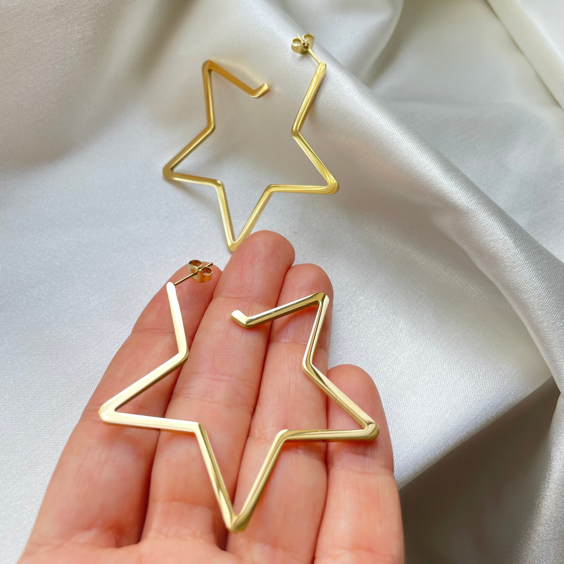 Stainless steel star hoops, 2.5 inches wide, push back hoop earring. Shown on hand to appreciate size. White silky background