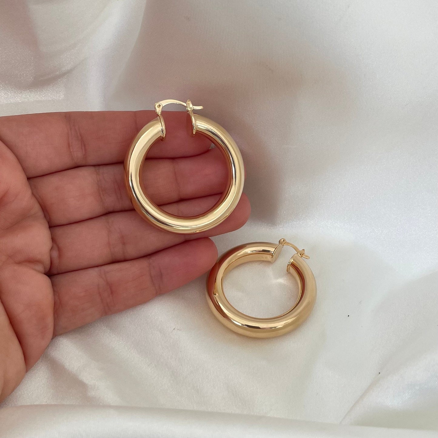 Chunky 18k gold filled hoop earrings, 6 mm thick and 1.7 inches diameter. Latch back closure. White silk background model is holding on hand the hoop earrings. 