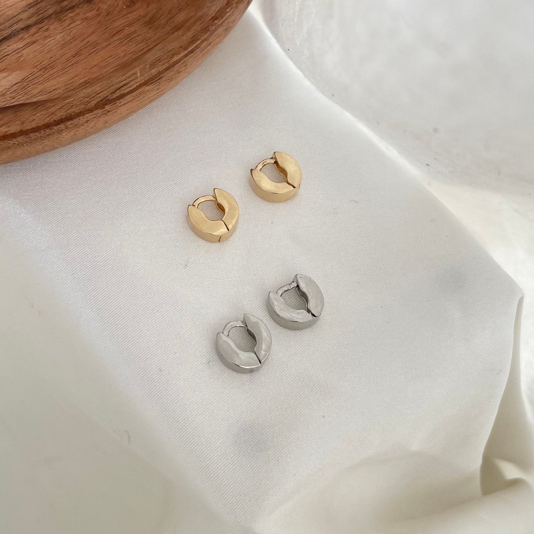 Two pairs of small huggie hoop earrings made of 18k gold filled or rhodium filled displayed on white silky fabric as background 