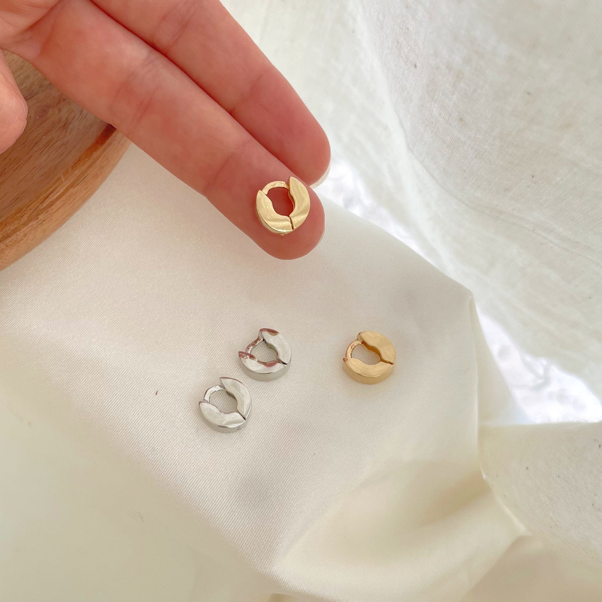 Holding on hand a small huggie hoop earring in 18k gold filled, at the back is the other gold hoop and a set of rhodium filled hoop earrings. White silky background