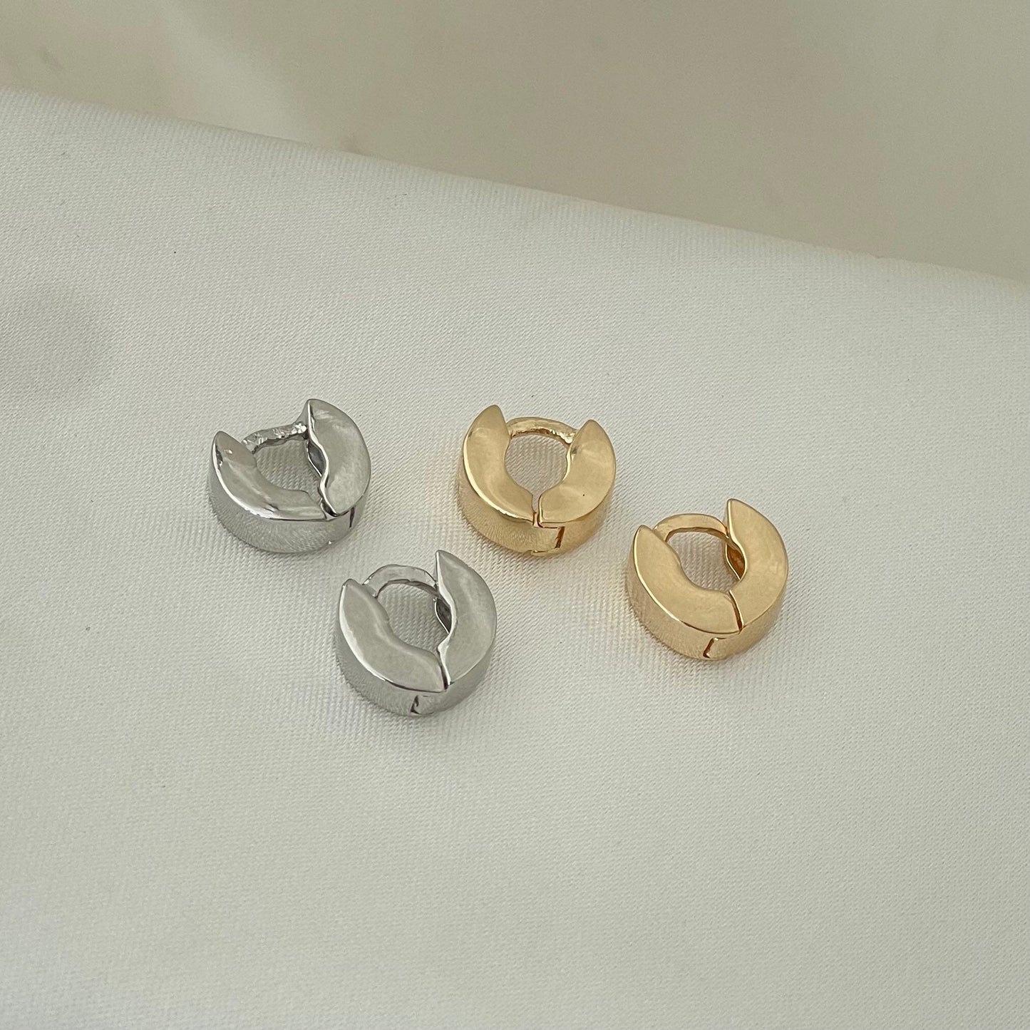 Two pairs of small huggie hoop earrings made of 18k gold filled or rhodium filled displayed on white silky fabric as background 