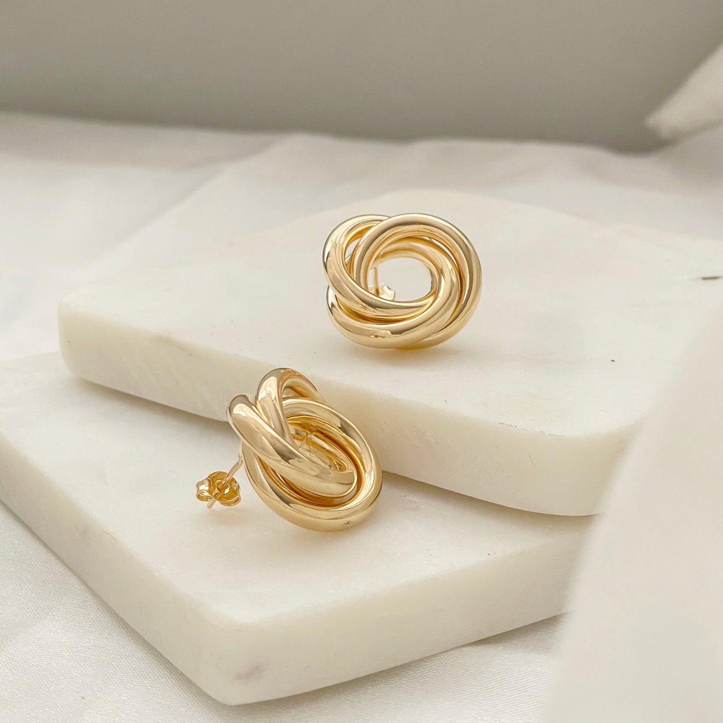 Front and side view of 18k gold filled knot stud earrings, chunky and chubby knot earrings displayed on marble and white silk background. Push back earrings 