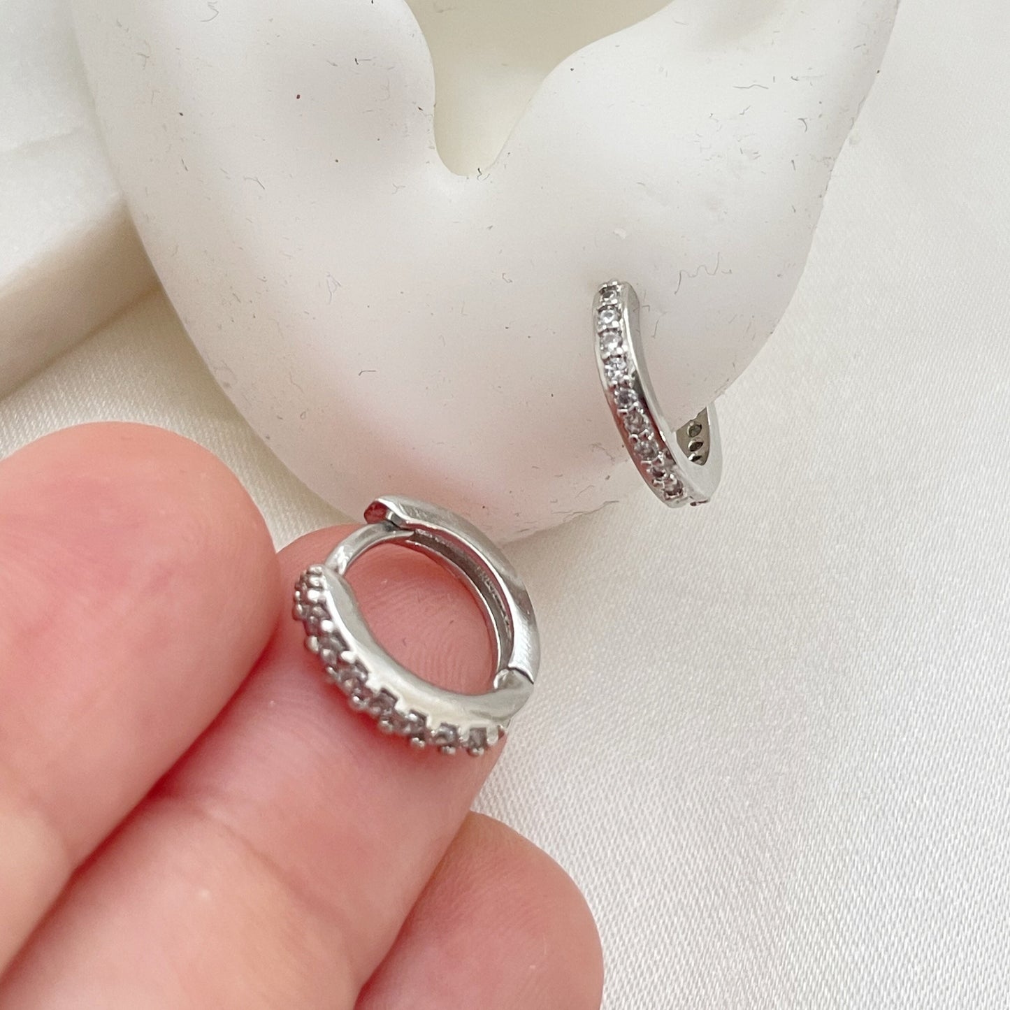 Cz huggie hoop earrings in Rhodium filled, displayed on silicone ear and white silky background