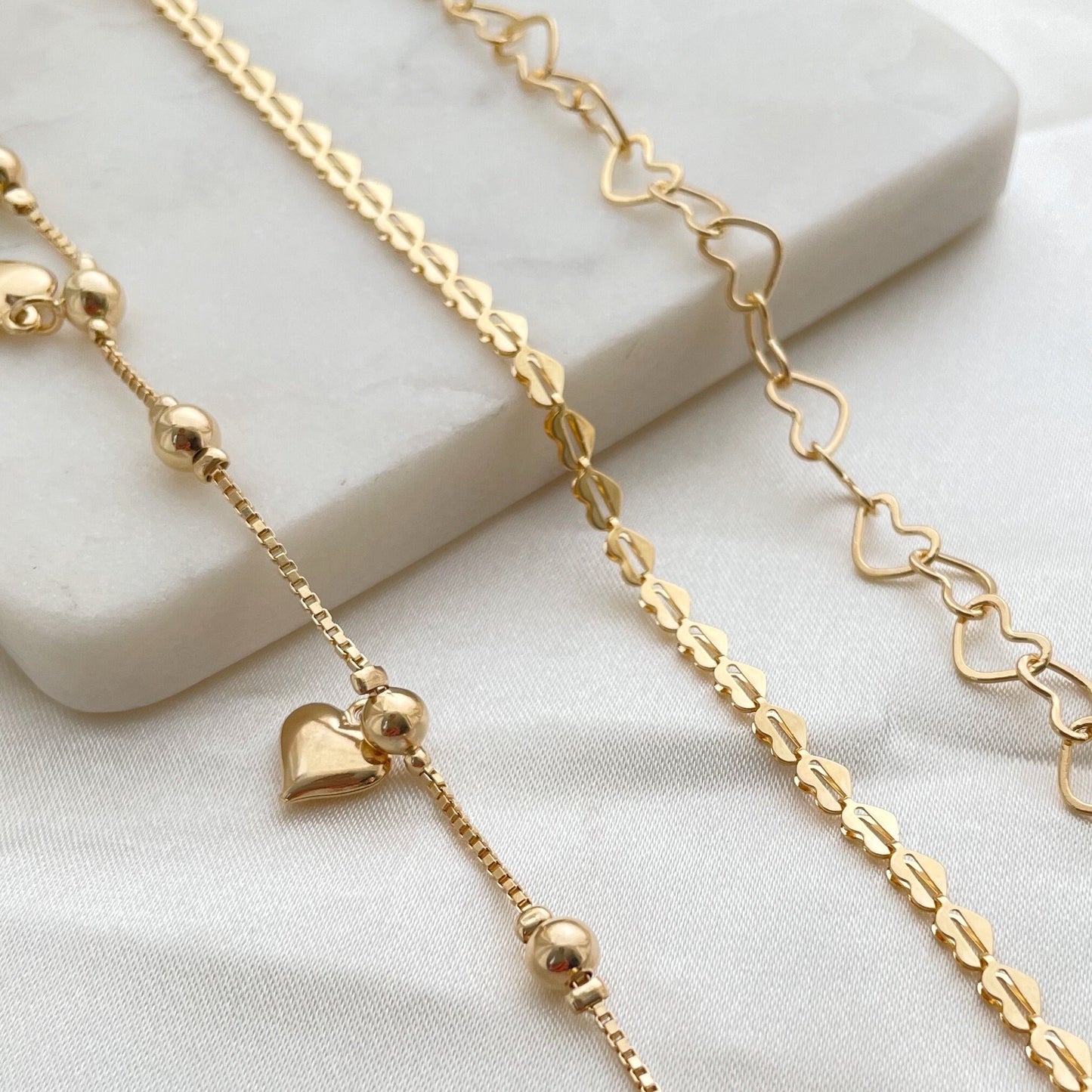 Set of 3 18k gold filled heart bracelets, 3 different styles: full heart bracelet, hollow heart link chain and dangle heart  bracelets. Displayed on a white marble tray. 