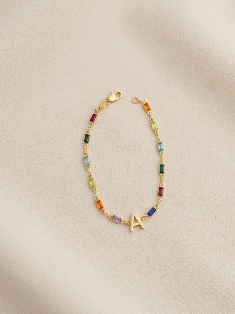 A delicate gold chain bracelet with small colorful gemstone beads and a gold initial 'A' charm, displayed on a soft beige fabric background.