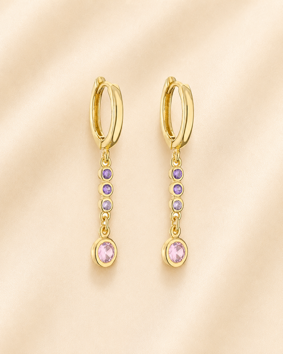 A pair of gold huggie hoop dangle earrings with small purple gemstones, displayed on a soft beige fabric background.