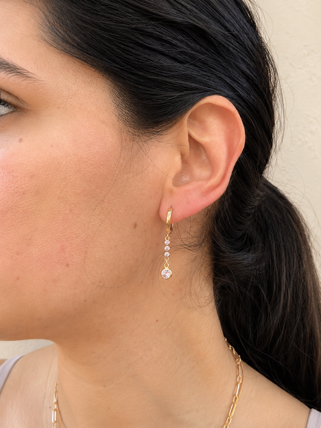 A close-up side profile of a woman wearing a delicate gold huggie hoop earring with a small drop of clear gemstones ending in a round clear stone, against a light beige wall.
