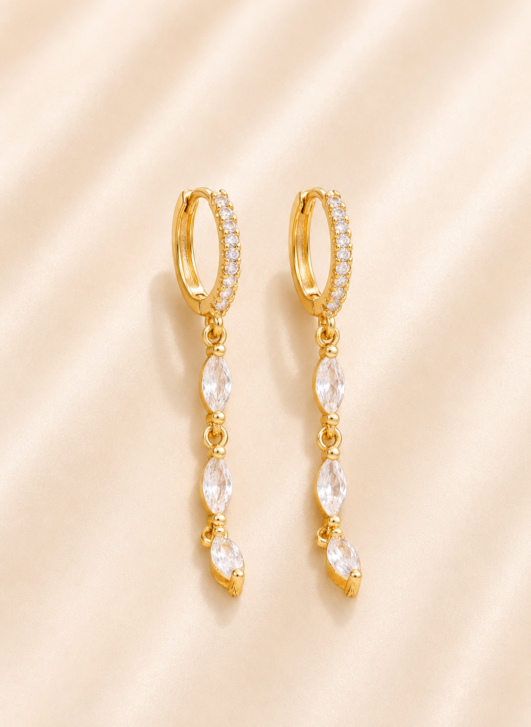 A pair of gold huggie hoop earrings with small clear stones along the top band and dangling marquise-cut clear stones, displayed on a soft beige satin background.