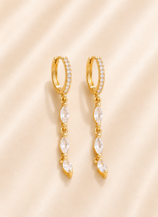 A pair of gold huggie hoop earrings with small clear stones along the top band and dangling marquise-cut clear stones, displayed on a soft beige satin background.