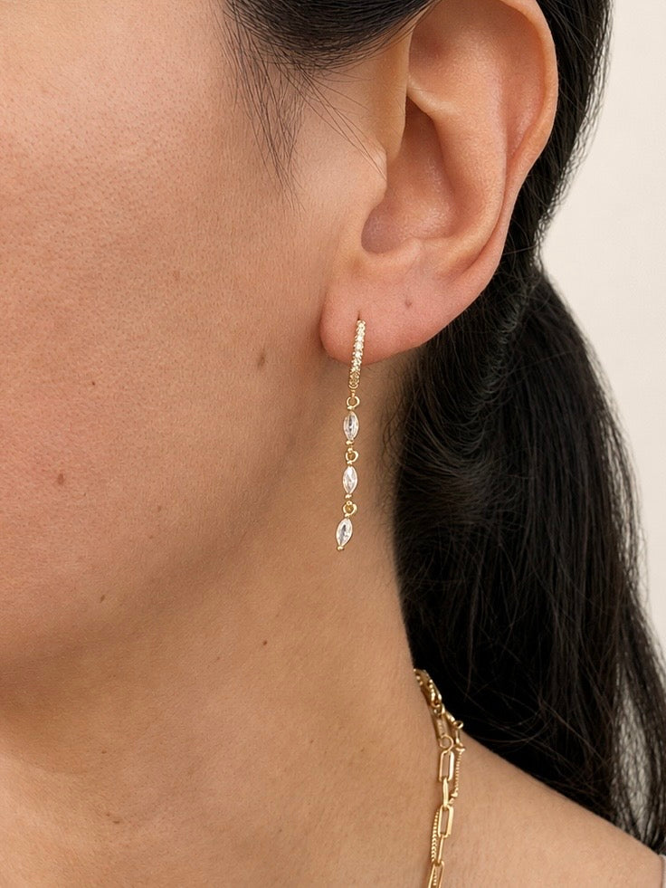 A close-up of a model's ear wearing a delicate gold huggie hoop earring with three dangling marquise-cut clear stones, paired with a gold chain necklace against a light beige background.
