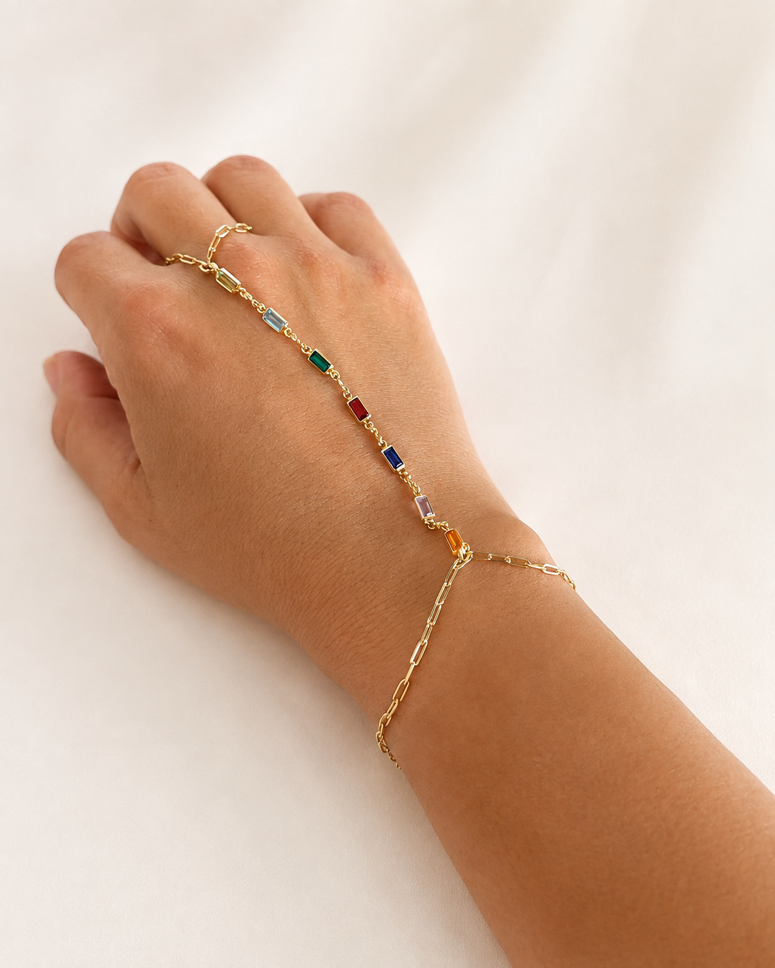 A delicate gold chain bracelet with small colorful gemstone accents, shown worn on a person's wrist and hand against a soft white background.