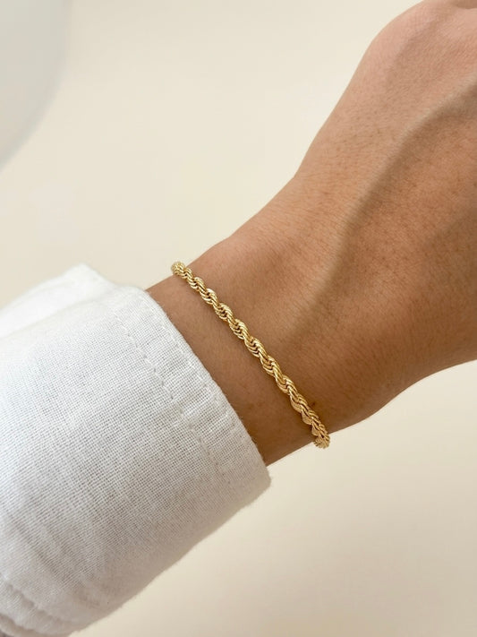 A delicate gold rope-style chain bracelet worn on a person's wrist, shown against a soft cream background with a white sleeve visible.