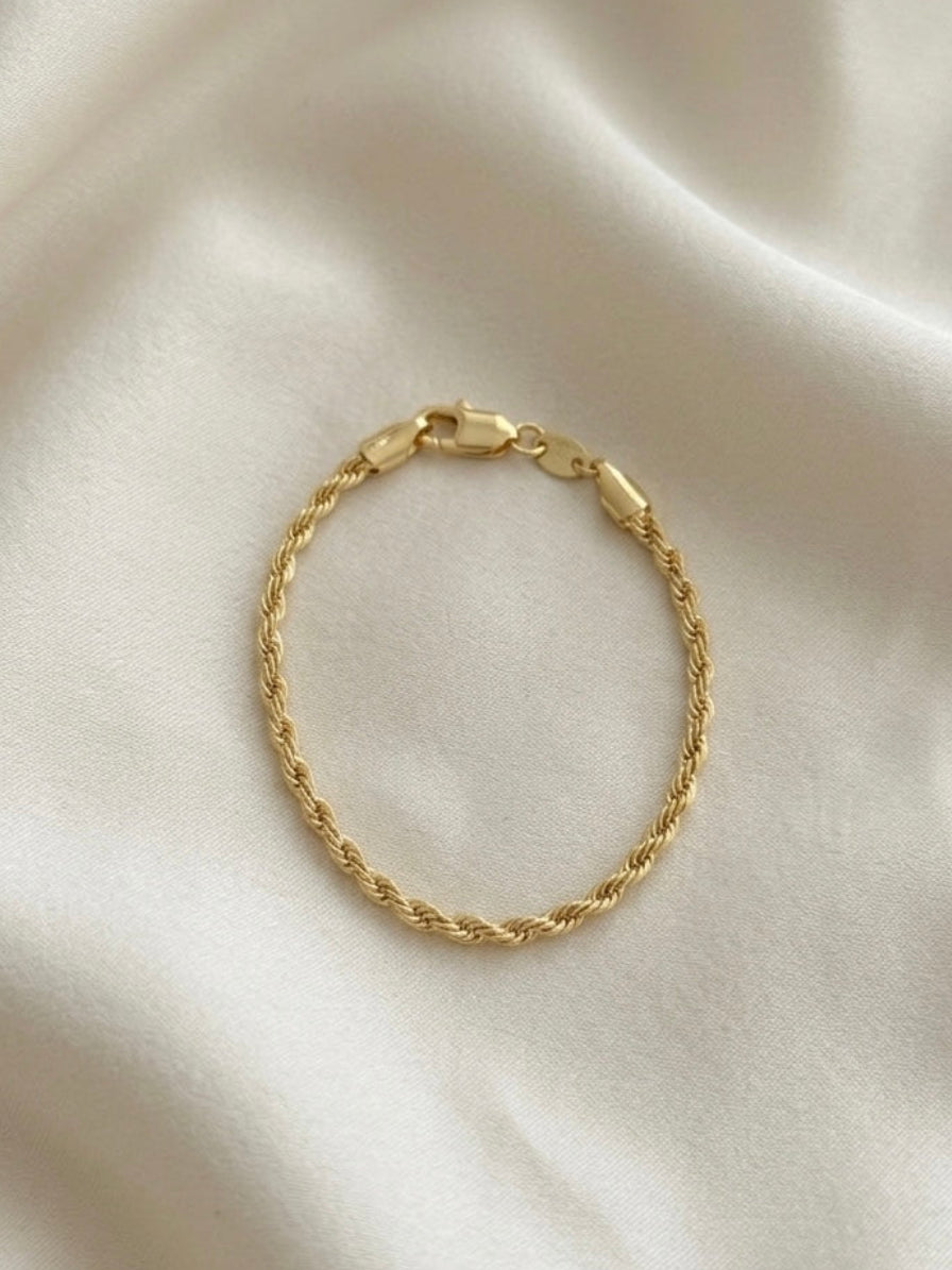 A delicate gold rope chain bracelet with a lobster clasp, displayed on a draped cream satin fabric background.