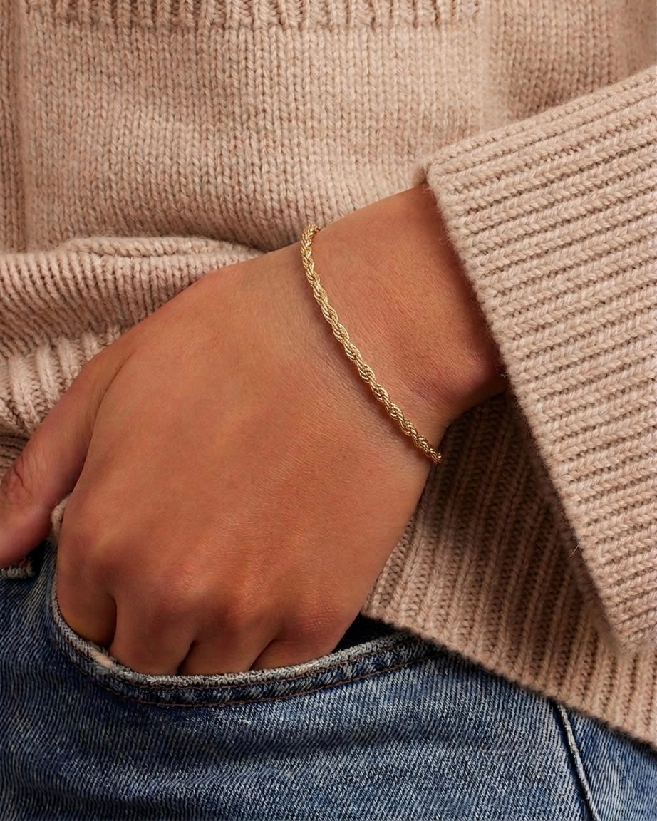 A delicate gold chain bracelet worn on a person's wrist, shown in a close-up lifestyle shot with a beige ribbed knit sweater and blue jeans.