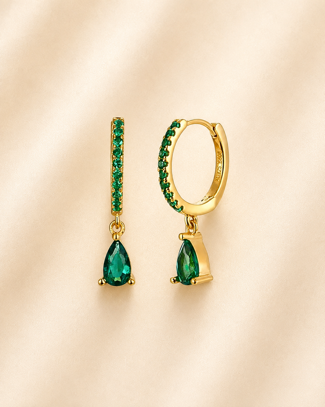 A pair of gold huggie hoop earrings with green gemstone detailing and teardrop-shaped green drops, displayed on a soft beige fabric background.