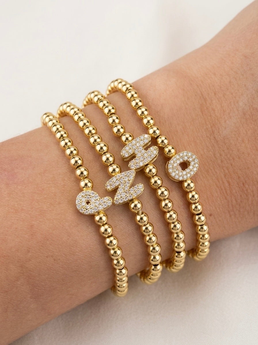 A close-up of a person's wrist wearing four gold beaded stretch bracelets, with letter charms in a pavé-set crystal design.