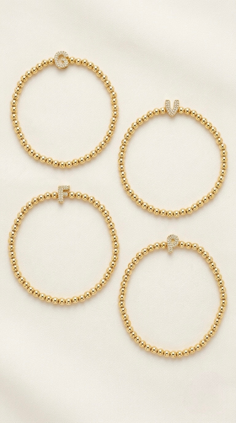 Four gold plated beaded bracelets, stretchy with small pave letter, arranged on a light cream fabric background.