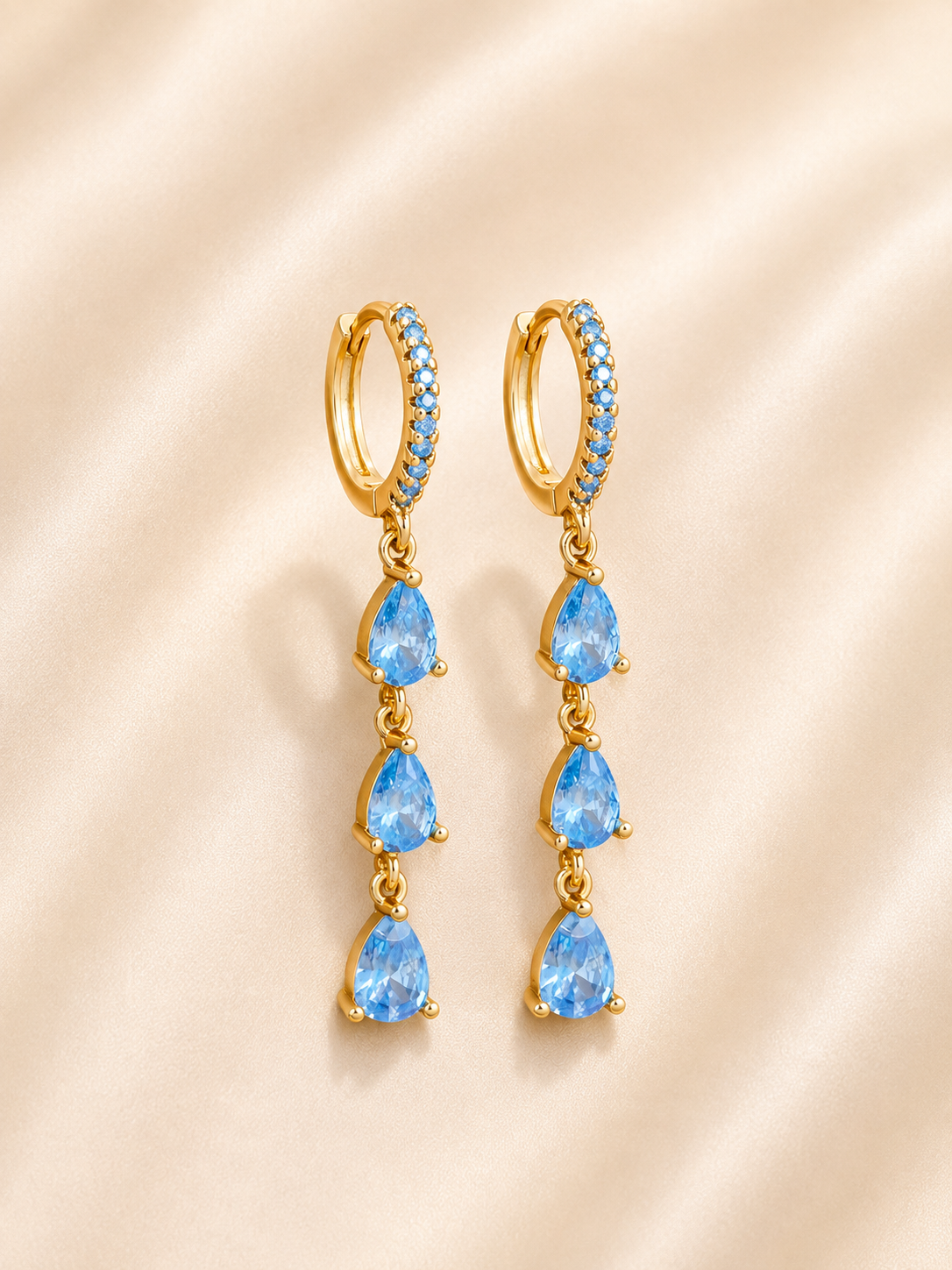 A pair of gold huggie hoop drop earrings featuring three blue teardrop-cut gemstones and a halo of small blue stones, displayed on a soft beige satin background.