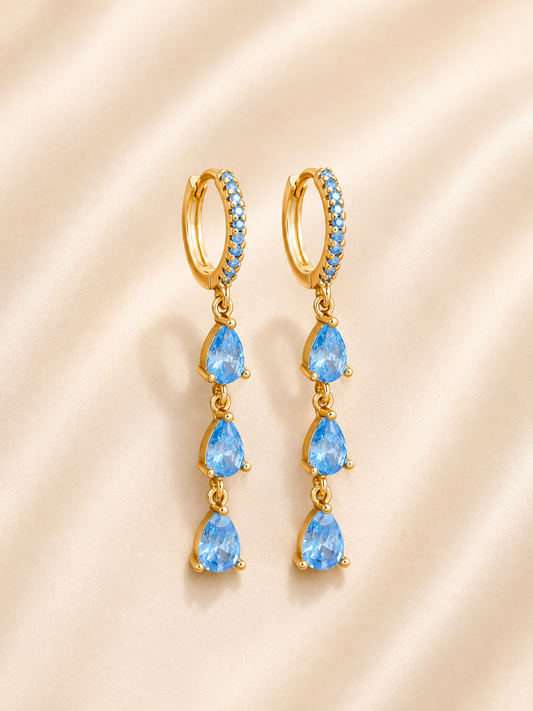 A pair of gold huggie hoop drop earrings featuring three blue teardrop-cut gemstones and a halo of small blue stones, displayed on a soft beige satin background.