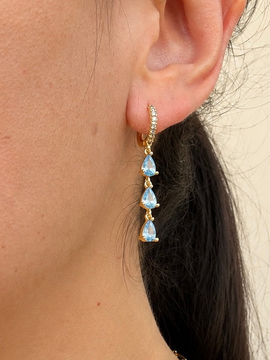 A close-up of a person's ear wearing a gold huggie hoop earring with a vertical drop of three teardrop-shaped light blue gemstones.